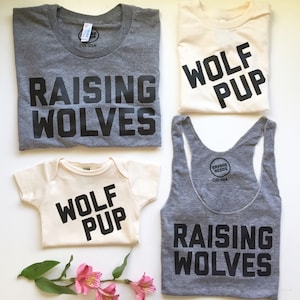 May include: A set of three shirts with the text "Raising Wolves" and "Wolf Pup" printed in black on a gray and cream background. The shirts are for adults and a baby.