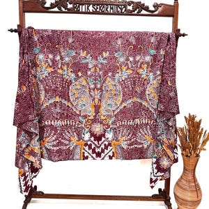 May include: A rectangular piece of fabric featuring a deep maroon background with intricate floral and peacock patterns in shades of yellow, white, and teal. The textile is draped over a wooden stand with the text BATIK SEKARMULYO carved at the top.