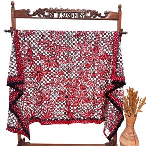 Può includere: Un pezzo di tessuto rettangolare con un motivo batik rosso, nero e bianco con disegni floreali e una griglia. Il tessuto è drappeggiato su un supporto in legno con la scritta BATIK SEKAR MULYO intagliata sulla traversa superiore. Un vaso intrecciato si trova accanto.