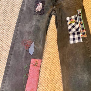 May include: Faded black denim jeans featuring red trim on the front pockets, a dark embroidered patch, a blue fabric teardrop, and two rectangular fabric patches in red gingham and navy blue plaid with floral embroidery and green stitching.