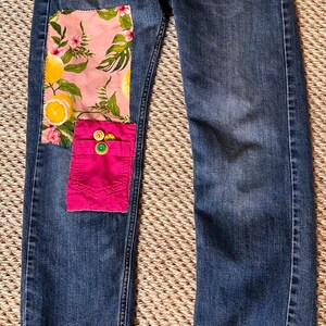 May include: Blue denim jeans with decorative patches. One patch features a pink floral and lemon print, and another is a bright pink pocket with colorful buttons. The jeans have green stitching and accents.