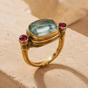 May include: A gold ring featuring a large, rectangular, light blue gemstone set in the center, flanked by two smaller, round, deep red gemstones on either side. The metal band has a textured, aged finish with detailed metalwork around the settings.