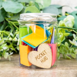 May include: A clear jar filled with colorful folded paper notes and small paper containers. A wooden heart with the words "POCKET HUG" is attached to the jar. The jar has a silver lid and is tied with a silver string.