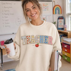 Puede incluir: Una sudadera de cuello redondo color crema con la palabra TEACHER en letras grandes de estilo bloque. Cada letra tiene un patrón único, incluyendo papel rayado, manzanas rojas, lápices amarillos, flores, cuadros vichy azules y lunares rosas.