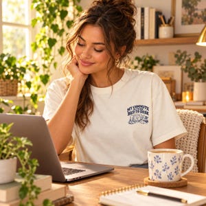 Puede incluir: Una camiseta de manga corta color blanco roto con un pequeño gráfico azul en el pecho izquierdo. El gráfico muestra un ordenador y un escritorio con el texto "MY COMMUTE IS FROM BED TO DESK" en una fuente azul oscuro y negrita.