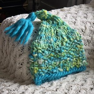 May include: A chunky knit beanie featuring a variegated blend of turquoise, lime green, and yellow yarn. The hat has a thick, solid turquoise braided brim and a matching turquoise yarn tassel attached to the pointed top of the textured crown.
