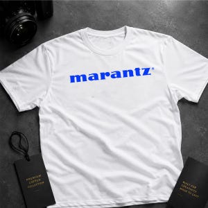 May include: White t-shirt featuring the blue word "marantz" across the chest. The shirt has a crew neck and short sleeves. Two black tags are present, one with "Premium Cotton Collection" and the other with "Built for Creativity Made to Last".