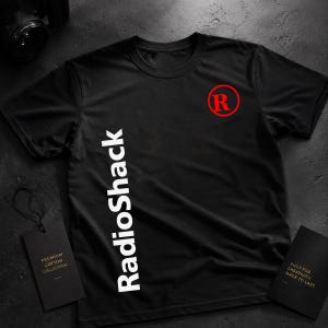 Pode incluir: Uma t-shirt preta de manga curta com o logótipo RadioShack na vertical em branco no lado esquerdo e um logótipo R vermelho num círculo no peito direito. A peça é acompanhada por duas etiquetas retangulares pretas com texto em dourado.