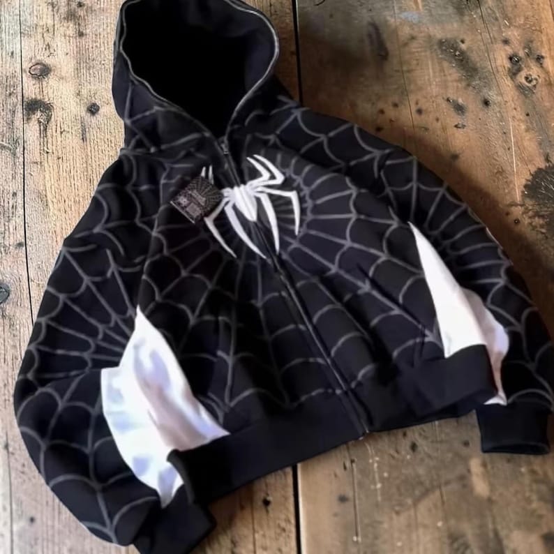 May include: A black zip-up hoodie featuring a white spider emblem on the chest and a grey spiderweb pattern across the fabric. The garment includes white side panels, a hood, and a black tag attached to the zip pull near the neckline.