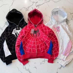 May include: Three zip-up hoodies in black, red, and white. Each hoodie features a spiderweb pattern across the fabric, a central spider graphic on the chest, and a full-length front zip. The black hoodie has white accents, the red hoodie has blue side panels, and the white hoodie has light pink side panels.