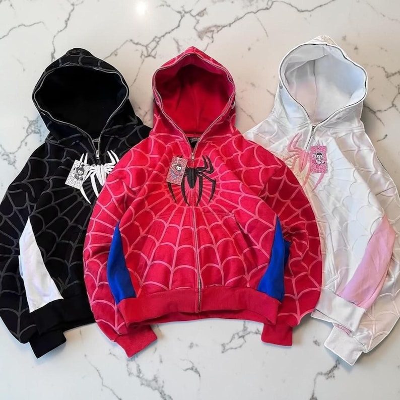 May include: Three zip-up hoodies in black, red, and white. Each hoodie features a spiderweb pattern across the fabric, a central spider graphic on the chest, and a full-length front zip. The black hoodie has white accents, the red hoodie has blue side panels, and the white hoodie has light pink side panels.