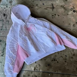 May include: A white zip-up hoodie featuring a raised, textured spiderweb pattern across the front and sleeves. The garment includes light pink side panels and a prominent pink spider graphic centred on the chest, with a hood and long sleeves.