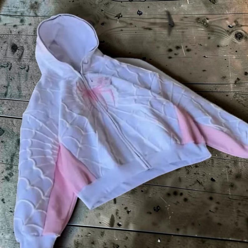 May include: A white zip-up hoodie featuring a raised, textured spiderweb pattern across the front and sleeves. The garment includes light pink side panels and a prominent pink spider graphic centred on the chest, with a hood and long sleeves.
