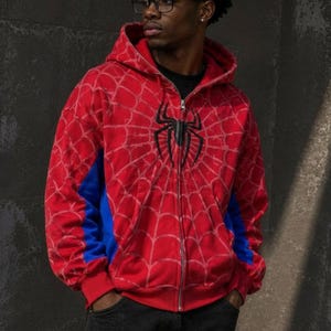 May include: A red zip-up hoodie featuring a black spider emblem on the chest and a white spiderweb pattern covering the front and sleeves. The garment includes blue side panels, a hood, and a full-length silver zip closure down the centre.