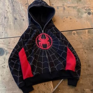May include: A black zip-up hoodie featuring a red spider emblem on the chest and a grey spiderweb pattern across the fabric. The garment includes a hood, long sleeves with red side panels, and a full-length silver-toned metal zipper closure.