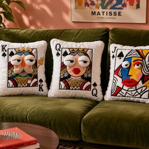 May include: Three square white plush throw pillows featuring embroidered playing card designs. The left pillow shows a King of Spades, the center shows a Queen of Spades, and the right shows a Jack of Spades, all with textured 3D facial features.