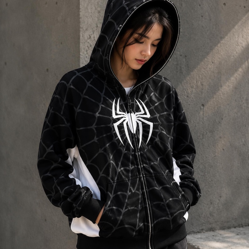 May include: A black zip-up hoodie featuring a white spider emblem on the chest and a repeating spider web pattern across the fabric. The garment includes a hood with a white inner lining, long sleeves with ribbed cuffs, and white side panels.