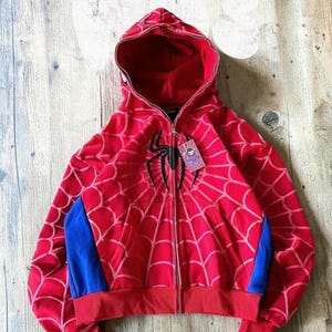 May include: A red zip-up hoodie featuring a black spider emblem on the chest and a web pattern across the fabric. The garment includes blue side panels, a hood, and a small rectangular tag attached to the zipper pull with a cartoon character.