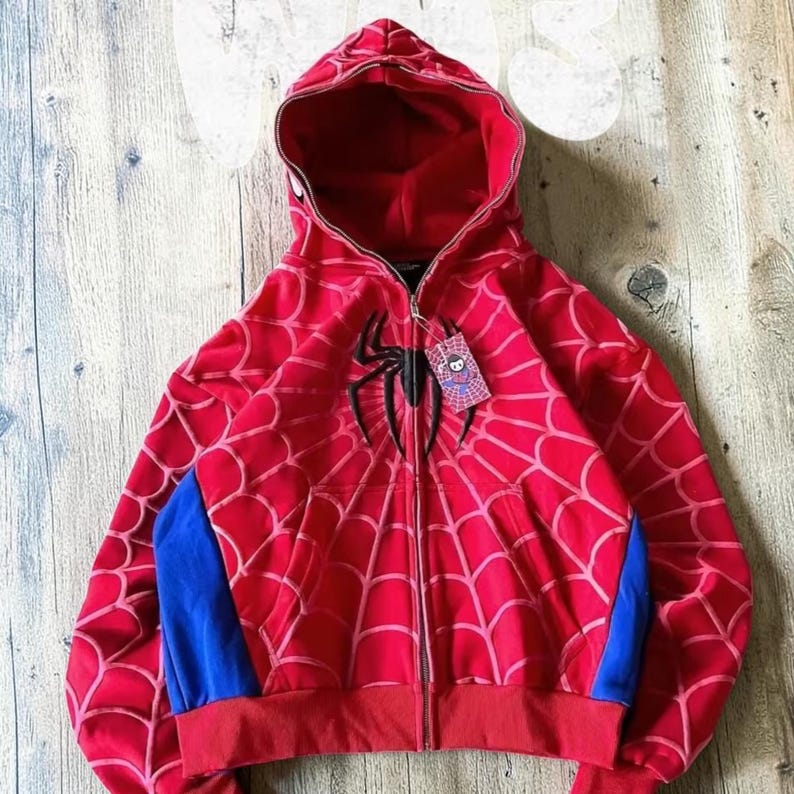 May include: A red zip-up hoodie featuring a black spider emblem on the chest and a web pattern across the fabric. The garment includes blue side panels, a hood, and a small rectangular tag attached to the zipper pull with a cartoon character.