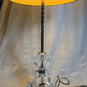 May include: A clear crystal table lamp with a cream-colored lampshade. The lamp features a faceted base, a diamond-shaped crystal accent, and a clear glass column with a dark central rod. The lampshade is pleated.