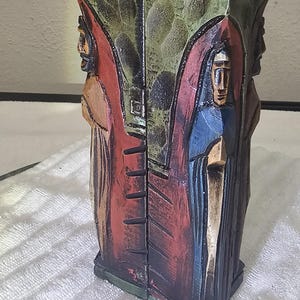 May include: A tall, vertical wooden sculpture featuring carved figures in relief. The piece has a peaked roof design, with a textured green upper section and figures painted in blue, red, and brown tones, set against a dark, carved background.