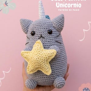 Puede incluir: Un peluche de gato tejido a ganchillo en color gris con un cuerno de unicornio blanco, sosteniendo una estrella tejida amarilla. El gato tiene ojos negros y bigotes bordados. El texto en la imagen dice Gato Pusheen Unicornio PATRÓN DE PAGO.