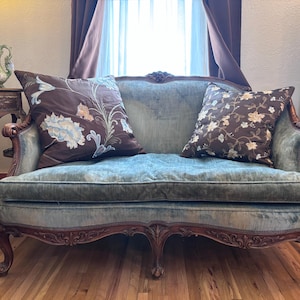 May include: A vintage blue velvet loveseat with ornate wooden detailing. Two decorative throw pillows with floral patterns in brown and cream tones rest on the seat. The sofa has curved legs and armrests, showcasing a classic design.