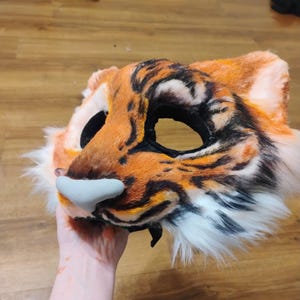 Puede incluir: Una m&aacute;scara de piel sint&eacute;tica con dise&ntilde;o de tigre en colores naranja, negro y blanco. La m&aacute;scara cuenta con grandes aberturas para los ojos, orejas puntiagudas y una pieza nasal esculpida en color gris claro. El patr&oacute;n imita rayas de tigre.