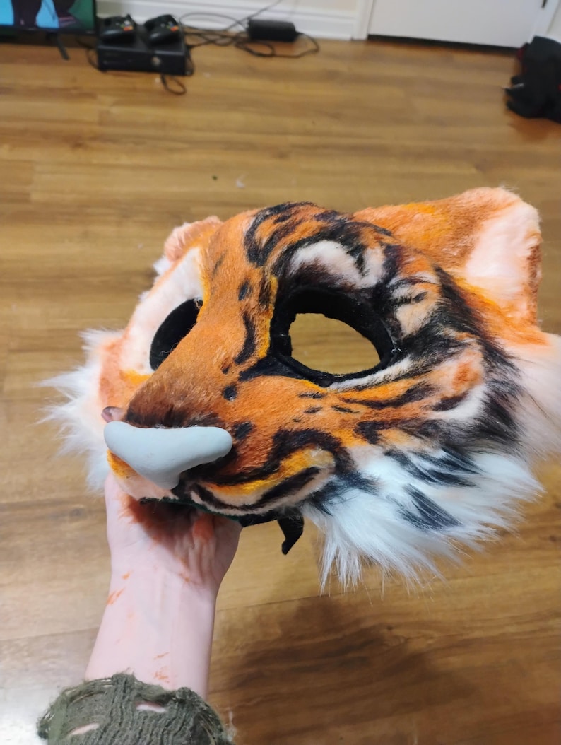Puede incluir: Una m&aacute;scara de piel sint&eacute;tica con dise&ntilde;o de tigre en colores naranja, negro y blanco. La m&aacute;scara cuenta con grandes aberturas para los ojos, orejas puntiagudas y una pieza nasal esculpida en color gris claro. El patr&oacute;n imita rayas de tigre.