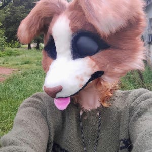 Puede incluir: Una m&aacute;scara de animal de piel sint&eacute;tica marr&oacute;n y blanca con grandes cuencas oculares negras, una nariz marr&oacute;n y una lengua rosa que sobresale. La m&aacute;scara se lleva con un su&eacute;ter de punto verde oliva con un patr&oacute;n negro y un collar con cruz de plata.