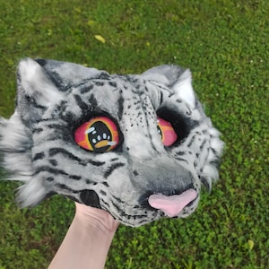 Puede incluir: Una m&aacute;scara de cabeza de animal de piel sint&eacute;tica con un patr&oacute;n de manchas grises y blancas que recuerda a un leopardo de las nieves. La m&aacute;scara tiene ojos grandes y expresivos con degradados naranja y rosa, pupilas negras y una nariz rosa claro.