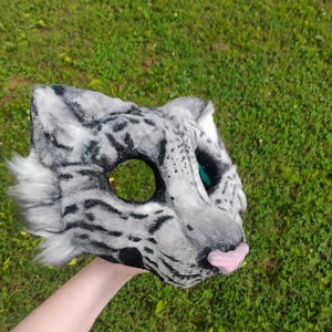 Puede incluir: Una m&aacute;scara de animal de piel sint&eacute;tica con un patr&oacute;n de manchas grises y blancas que recuerda a un leopardo de las nieves. La m&aacute;scara tiene orejas puntiagudas, cuencas oculares negras, una nariz rosa y largos mechones de pelo blanco a los lados.