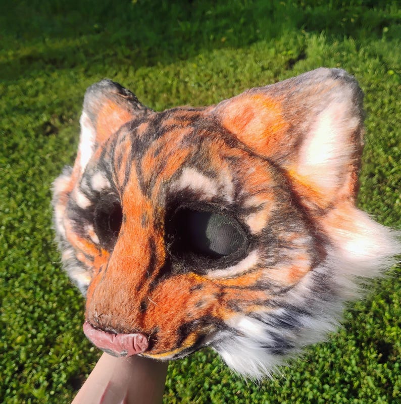 Puede incluir: Una m&aacute;scara de animal con dise&ntilde;o de tigre hecha de piel sint&eacute;tica naranja, negra y blanca con orejas puntiagudas, aberturas oscuras para los ojos y una nariz rosa. La m&aacute;scara muestra un patr&oacute;n de rayas realista con acentos de piel blanca.