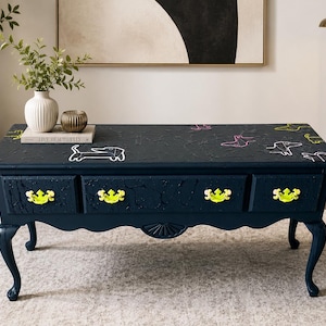 May include: A rectangular dark navy blue coffee table with four cabriole legs and four drawers featuring bright neon yellow hardware. The tabletop surface has a textured finish and is decorated with white, pink, and yellow line art of dogs.