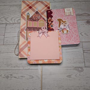 May include: A set of three paper stationery items including a tall rectangular card with a house illustration, a pink notepad with a bow, and a pink notebook featuring a squirrel graphic and the text Fall, all decorated with plaid and floral patterns.