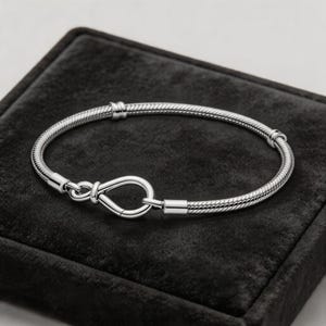 Könnte beinhalten: Ein silbernes Armband mit Schlangenkettendesign und einem Unendlichkeitszeichen-Verschluss. Das Armband wird auf einer schwarzen Samtoberfläche präsentiert, die seine polierte Oberfläche und die komplizierten Details hervorhebt. Ein stilvolles Schmuckstück.