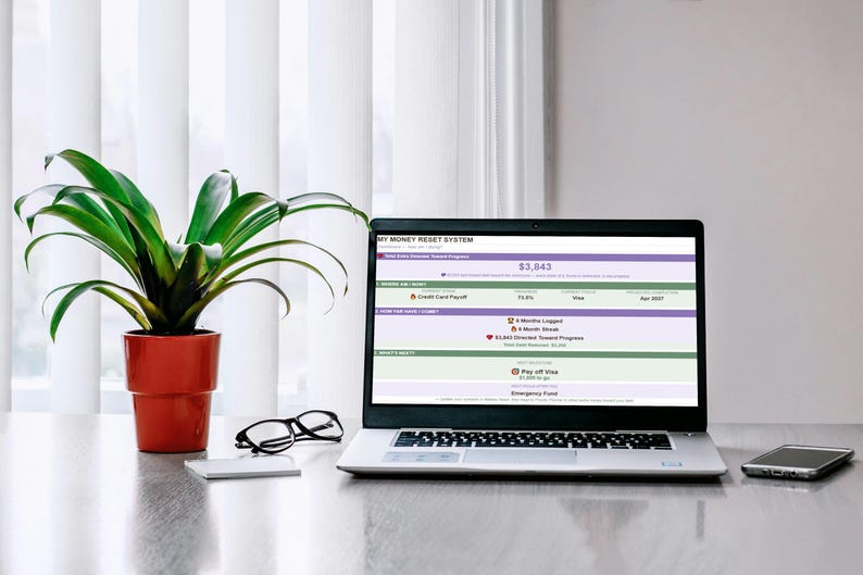 May include: A silver laptop displaying a My Money Reset System dashboard with financial progress bars, a green potted plant in an orange pot, black-rimmed eyeglasses, and a smartphone. The screen shows data including a $3,843 total and credit card payoff goals.