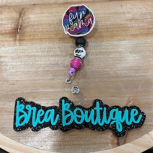 May include: A black and turquoise glitter name tag with the text "Brea Boutique". A colorful paw print with the text "Fur Mama" is attached to a black and white beaded lanyard.
