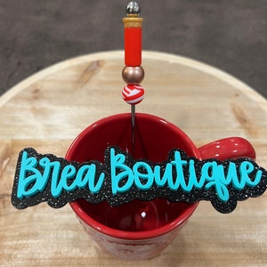 May include: A red mug with the words "Brea Boutique" in turquoise lettering on a black background. A red and gold beaded stir stick is in the mug.