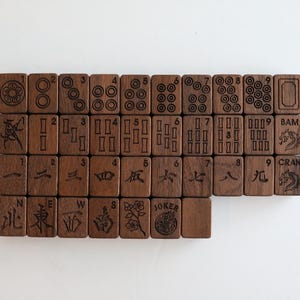 Pu&ograve; includere: Un set di tessere rettangolari da mahjong in legno marrone scuro disposte in una griglia. Le tessere presentano incisioni nere che includono numeri, cerchi, motivi di bamb&ugrave;, caratteri cinesi, punti cardinali, un fiore e un drago con testo.