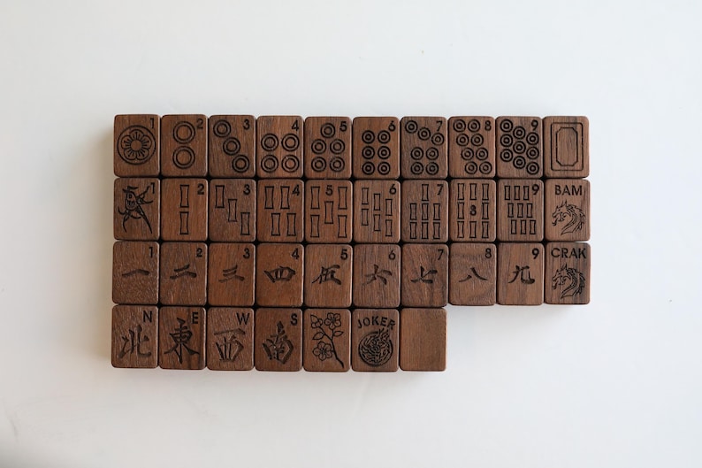 Pu&ograve; includere: Un set di tessere rettangolari da mahjong in legno marrone scuro disposte in una griglia. Le tessere presentano incisioni nere che includono numeri, cerchi, motivi di bamb&ugrave;, caratteri cinesi, punti cardinali, un fiore e un drago con testo.
