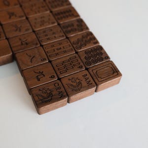 Pu&ograve; includere: Un set di tessere rettangolari da mahjong in legno marrone scuro disposte in file. Ogni tessera presenta disegni incisi al laser tra cui caratteri cinesi, cifre numeriche, motivi geometrici, cerchi e illustrazioni di draghi con il testo CRAK e BAM.