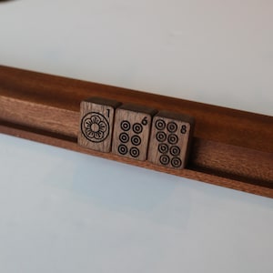 Pu&ograve; includere: Tre tessere rettangolari da mahjong in legno marrone scuro appoggiate su un supporto in legno. La prima tessera presenta un simbolo floreale e il numero 1. La seconda tessera mostra sei motivi circolari e il numero 6. La terza tessera mostra otto motivi circolari e il numero 8.