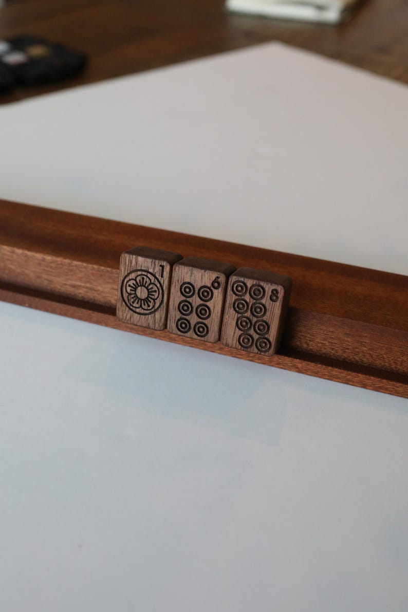 Pu&ograve; includere: Tre tessere rettangolari da mahjong in legno marrone scuro appoggiate su un supporto in legno. La prima tessera presenta un simbolo floreale e il numero 1. La seconda tessera mostra sei motivi circolari e il numero 6. La terza tessera mostra otto motivi circolari e il numero 8.
