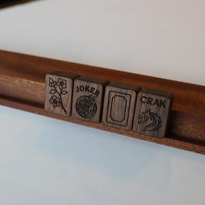 Pu&ograve; includere: Quattro tessere rettangolari da mahjong in legno scuro poggiate su un supporto in legno. Le tessere presentano disegni incisi: un fiore, la parola JOKER con un emblema circolare, un bordo rettangolare vuoto e la parola CRAK con un drago.