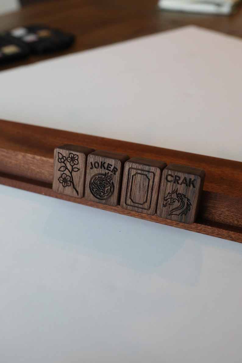 Pu&ograve; includere: Quattro tessere rettangolari da mahjong in legno scuro poggiate su un supporto in legno. Le tessere presentano disegni incisi: un fiore, la parola JOKER con un emblema circolare, un bordo rettangolare vuoto e la parola CRAK con un drago.