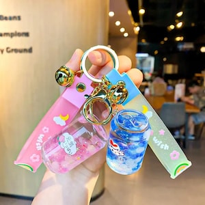 May include: A keychain set featuring two miniature clear plastic bottles filled with colorful beads, attached to a gold-tone metal ring. One bottle is pink with Japanese text, the other is blue with the text "pass". Two long, glittery silicone straps in pink and light blue accompany the charms, each printed with the word "wish", clouds, stars, and flowers.