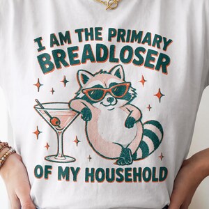 May include: A white t-shirt featuring a graphic of a raccoon wearing orange sunglasses, leaning against a martini glass with an olive. The text reads I AM THE PRIMARY BREADLOSER OF MY HOUSEHOLD in dark green and orange lettering with star accents.