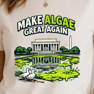 May include: A cream-coloured short-sleeve t-shirt featuring a graphic print of the Lincoln Memorial and Washington Monument with a green algae-covered reflecting pool, two white ducks, and the text "MAKE ALGAE GREAT AGAIN" in black and green.