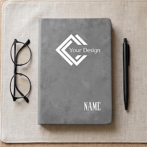 May include: A gray notebook featuring a white geometric logo with the text Your Design and the word NAME printed below it. To the left of the notebook are black-rimmed glasses, and to the right is a black pen, all arranged on a flat surface.